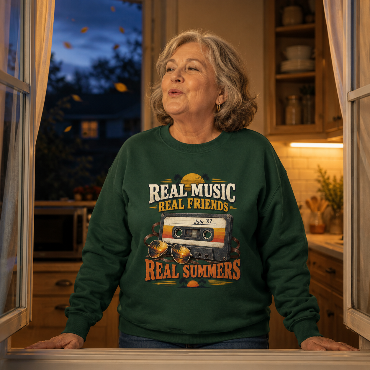 believable older woman in her early 60s with a fuller build and a calm expression wearing the "Real Music. Real Friends July '87 Real Summers" vintage sweatshirt as her outer layer, long sleeves clearly visible, on a cool evening, standing at an open kitchen window, humming an old summer tune while the cool air comes in, hands resting on the sill