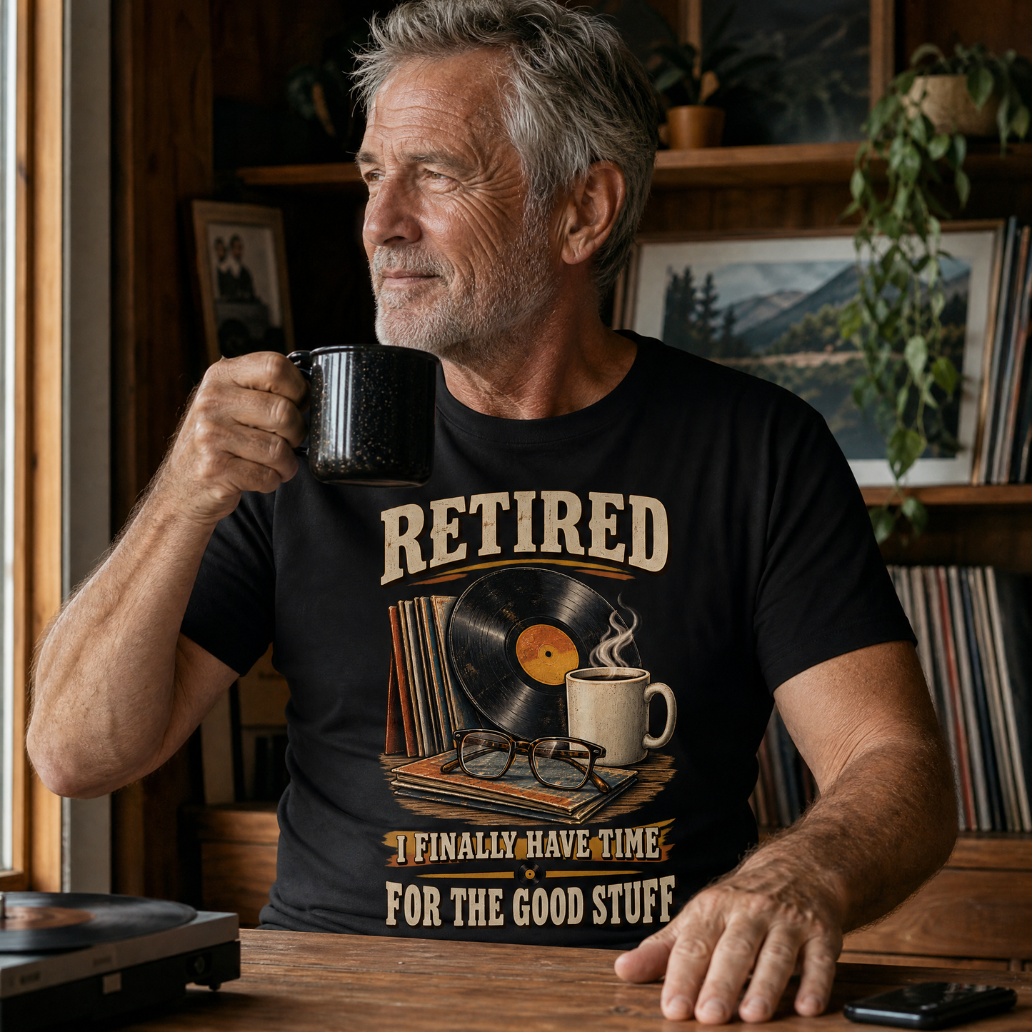 A man in a rustic room wearing a black graphic t-shirt that reads 'RETIRED I FINALLY HAVE TIME FOR THE GOOD STUFF' with a vinyl record, mug, and glasses illustration. He is holding a black coffee mug.