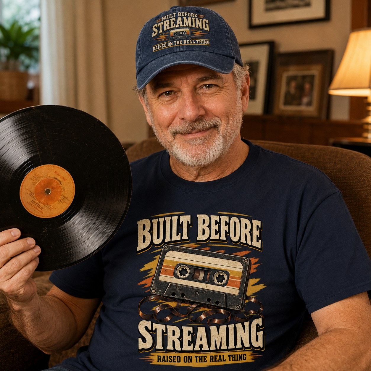 A smiling older man wearing a navy blue graphic T-shirt and matching baseball cap sits indoors while holding a black vinyl record. The shirt and cap both feature the text 'BUILT BEFORE STREAMING' and 'RAISED ON THE REAL THING' with a cassette graphic.