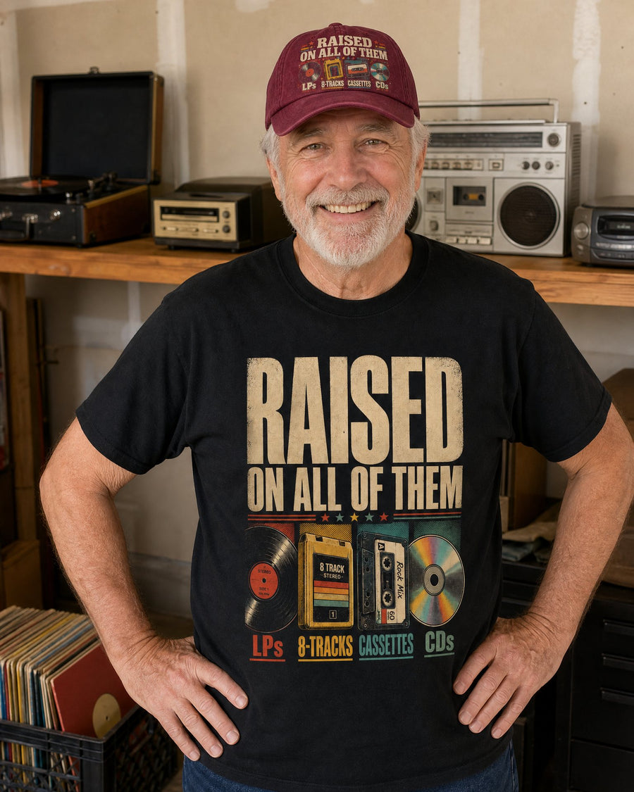Man in the Raised On All Of Them black tee and wine red cap in his garage, a record player, 8-track deck, boom box and CD player on the shelf behind