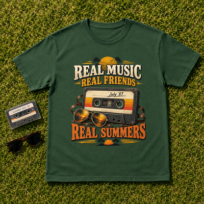 A dark green graphic t-shirt laid flat on green grass, styled with a vintage cassette tape, sunglasses, and a small cassette tape prop. The t-shirt features a retro graphic with the text 'REAL MUSIC REAL FRIENDS' and 'REAL SUMMERS' surrounding a cassette tape illustration labeled 'July 27'.