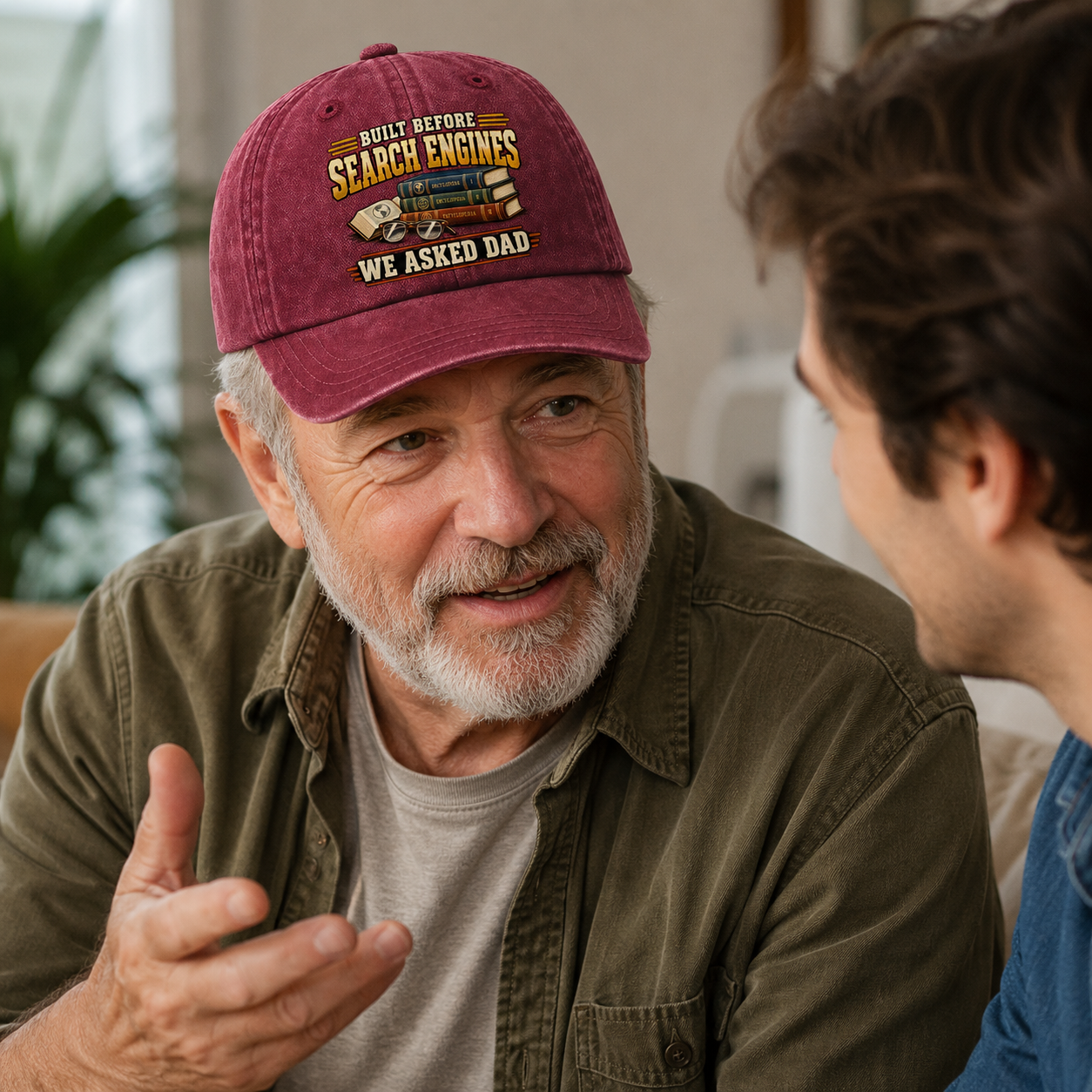 An older man wearing a maroon baseball cap with the text 'BUILT BEFORE SEARCH ENGINES WE ASKED DAD' and a graphic of a search engine, talking to a younger man in a softly lit indoor setting.