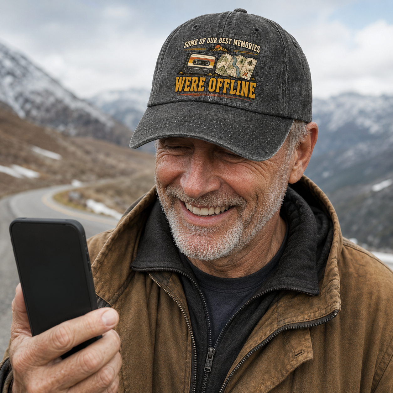 A smiling man outdoors in a mountain setting wears a dark gray baseball cap with a yellow and white graphic that reads 'SOME OF OUR BEST MEMORIES WE'RE OFFLINE' while holding a black smartphone.