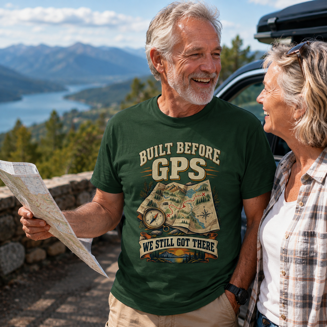 A smiling older man wearing a dark green graphic t-shirt stands outdoors next to a woman, holding a map in front of a car with a scenic mountain lake in the background. The t-shirt features a vintage-style map graphic and the text 'BUILT BEFORE GPS WE STILL GOT THERE'.