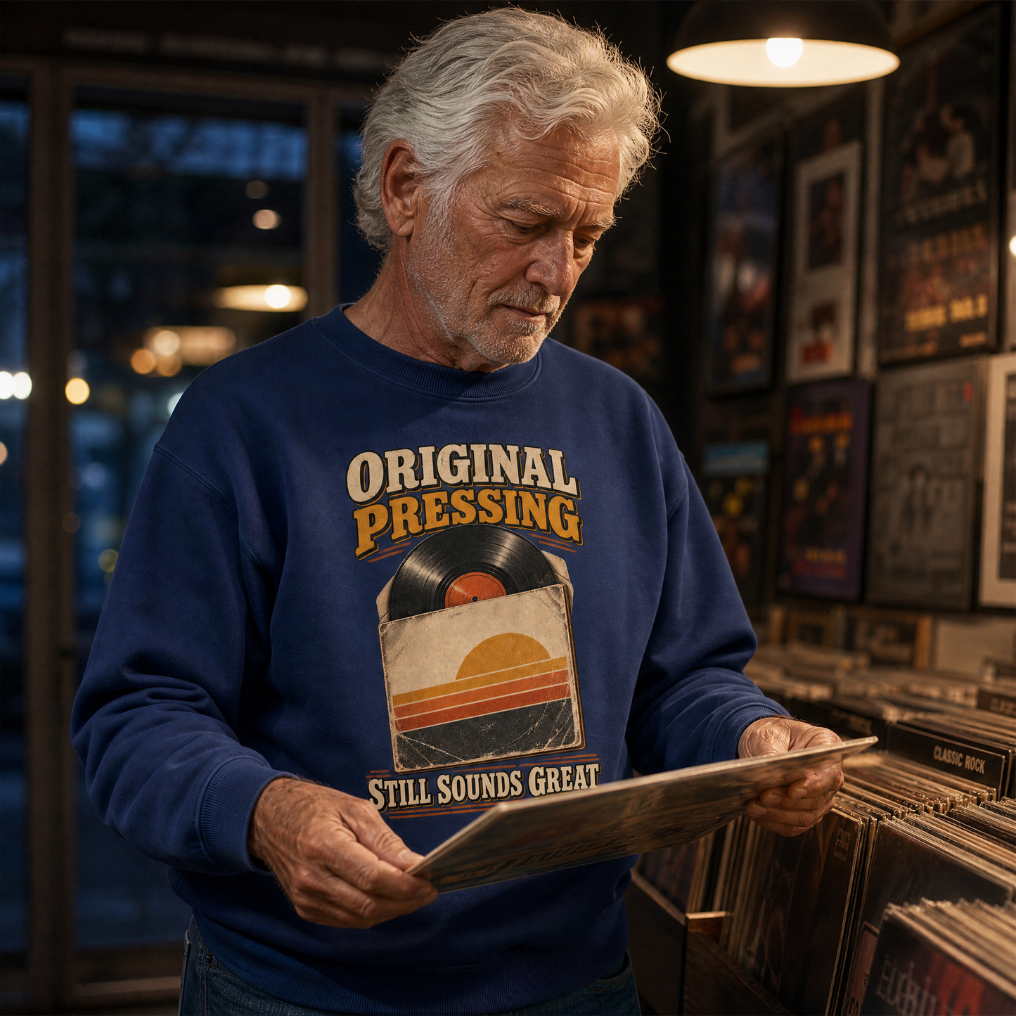 An older man with gray hair stands in a record store, holding a vinyl record sleeve. He is wearing a navy blue crewneck sweatshirt with a retro graphic that reads 'ORIGINAL PRESSING' and 'STILL SOUNDS GREAT' around a vinyl record and sunset illustration.