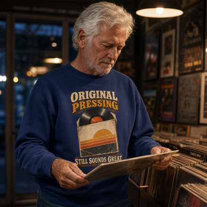 An older man with gray hair stands in a record store, holding a vinyl record sleeve. He is wearing a navy blue crewneck sweatshirt with a retro graphic that reads 'ORIGINAL PRESSING' and 'STILL SOUNDS GREAT' around a vinyl record and sunset illustration.