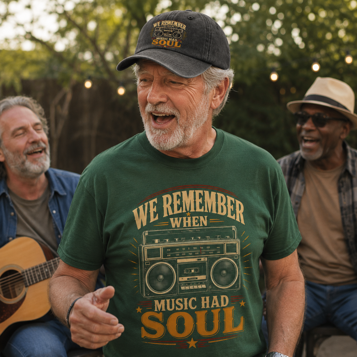 A man outdoors wearing a dark green graphic T-shirt that reads 'WE REMEMBER WHEN MUSIC HAD SOUL' with a vintage boombox illustration, along with a dark baseball cap. Two other men are seated in the background, one holding a guitar, in a softly lit outdoor setting with trees and string lights.