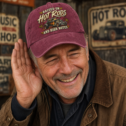 A smiling older man wearing a maroon baseball cap with a hot rod graphic and the text 'RAISED ON HOT RODS AND HIGH NOTES,' posed in front of vintage-style shop signs.