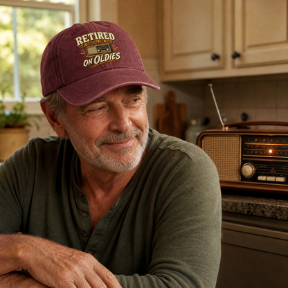 A smiling older man wearing a maroon baseball cap with 'RETIRED ON OLDIES' embroidered on the front, leaning in a warm kitchen setting with wooden cabinets and a vintage radio in the background.