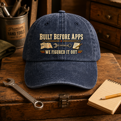 A blue distressed baseball cap with a vintage-style graphic reading 'BUILT BEFORE APPS WE FIGURED IT OUT' and tool illustrations, displayed on a wooden workbench with wrenches, a notebook, and a metal tin of hand tools.