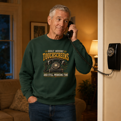 An older man wearing a dark green crewneck sweatshirt with a retro graphic that reads 'BUILT BEFORE TOUCHSCREENS AND STILL WORKING FINE' while holding a vintage black rotary phone in a warmly lit living room.