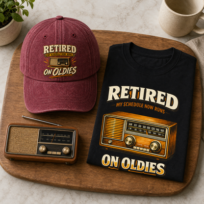 A folded black graphic T-shirt and a maroon baseball cap both printed with the phrase 'RETIRED ON OLDIES' and a vintage radio design, arranged on a wooden board with a small radio prop, mug, and plant.