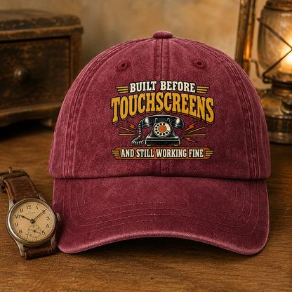 A maroon baseball cap with a vintage-style embroidered graphic reading 'BUILT BEFORE TOUCHSCREENS AND STILL WORKING FINE' around a retro telephone illustration. The cap is photographed on a wooden surface with a wristwatch and warm-toned background props, including a lantern.