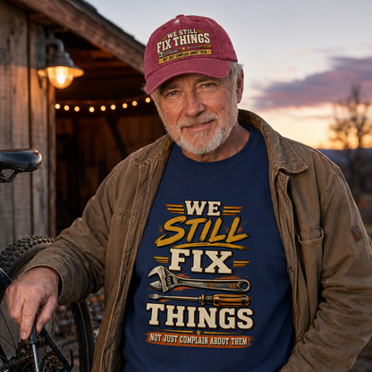 An older man wearing a maroon cap and a brown jacket stands outdoors at dusk in front of a wooden barn, holding a bicycle handlebar. He wears a navy graphic T-shirt with the slogan 'WE STILL FIX THINGS NOT JUST COMPLAIN ABOUT THEM' and wrench illustrations.