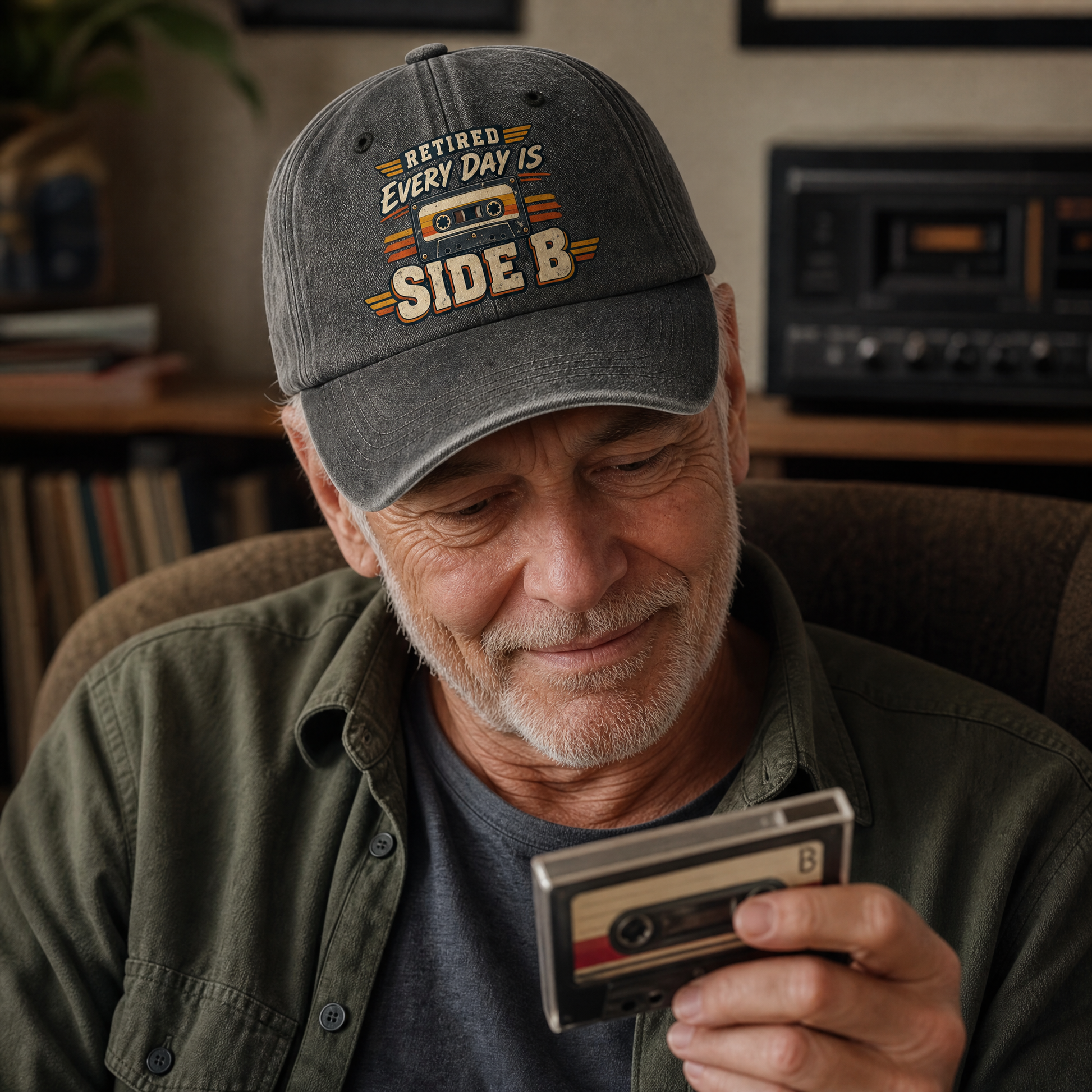 A smiling older man wearing a gray baseball cap with the text 'RETIREED EVERY DAY IS SIDE B' in yellow, orange, and white, holding a cassette tape recorder in a cozy indoor setting.