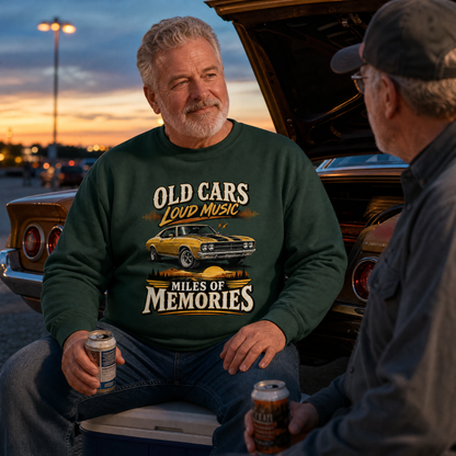 A man wearing a dark green crewneck sweatshirt with a graphic of a yellow muscle car and the text 'OLD CARS LOUD MUSIC MILES OF MEMORIES' sits in front of the open trunk of a classic car, holding a beer can in a outdoor setting at sunset.
