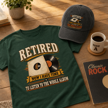 A dark green graphic T-shirt laid on a wooden surface, alongside a matching dark gray baseball cap, a white coffee mug, and a record-style book cover. The shirt and cap both feature a vintage-style print reading “RETIRED NOW I HAVE TIME TO LISTEN TO THE WHOLE ALBUM” with a vinyl record illustration.