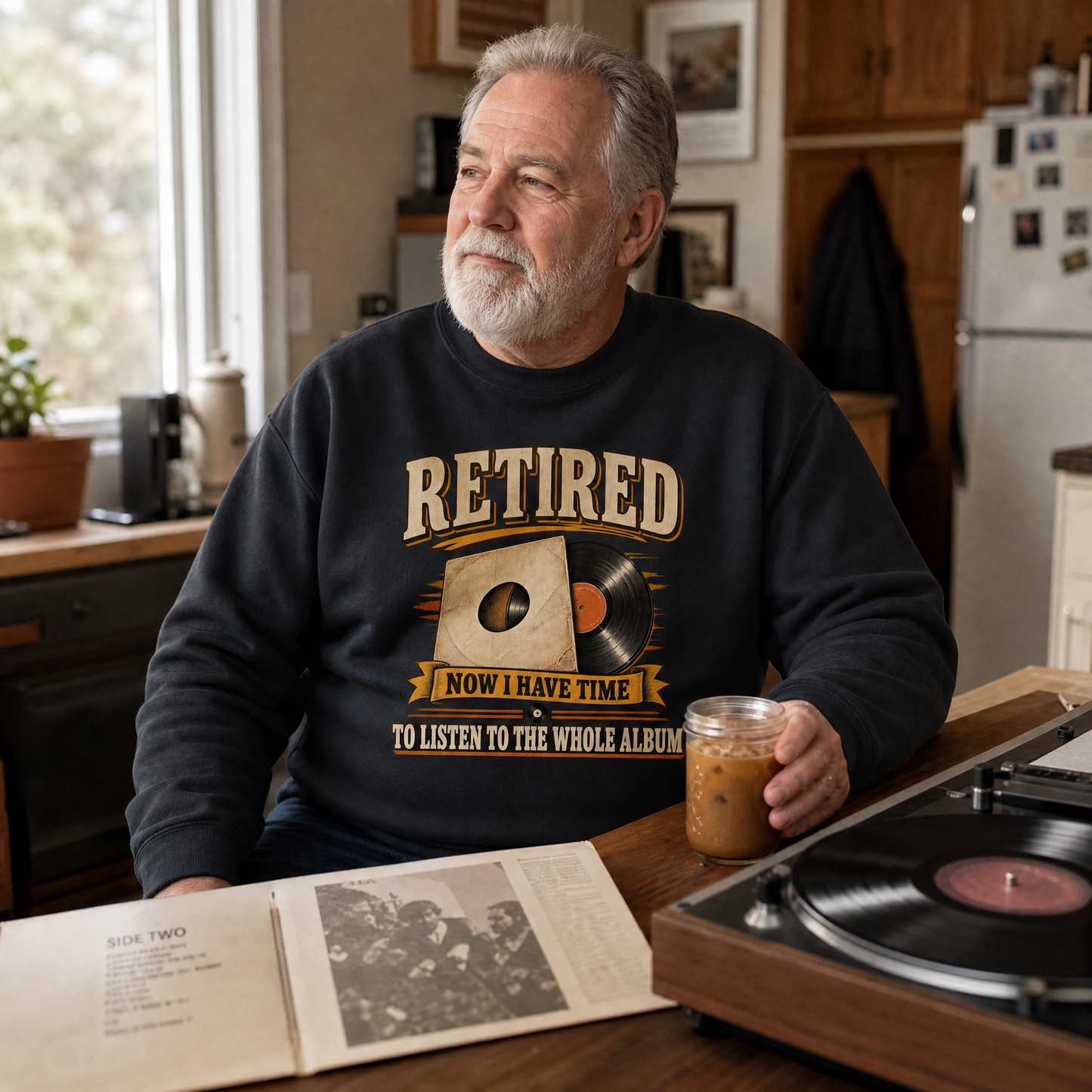 An older man with a gray beard sits at a wooden table in a kitchen setting, wearing a black crewneck sweatshirt with a vintage-style graphic that reads 'RETIRED NOW I HAVE TIME TO LISTEN TO THE WHOLE ALBUM'. He is holding a glass jar of iced coffee next to a wooden turntable with a vinyl record.