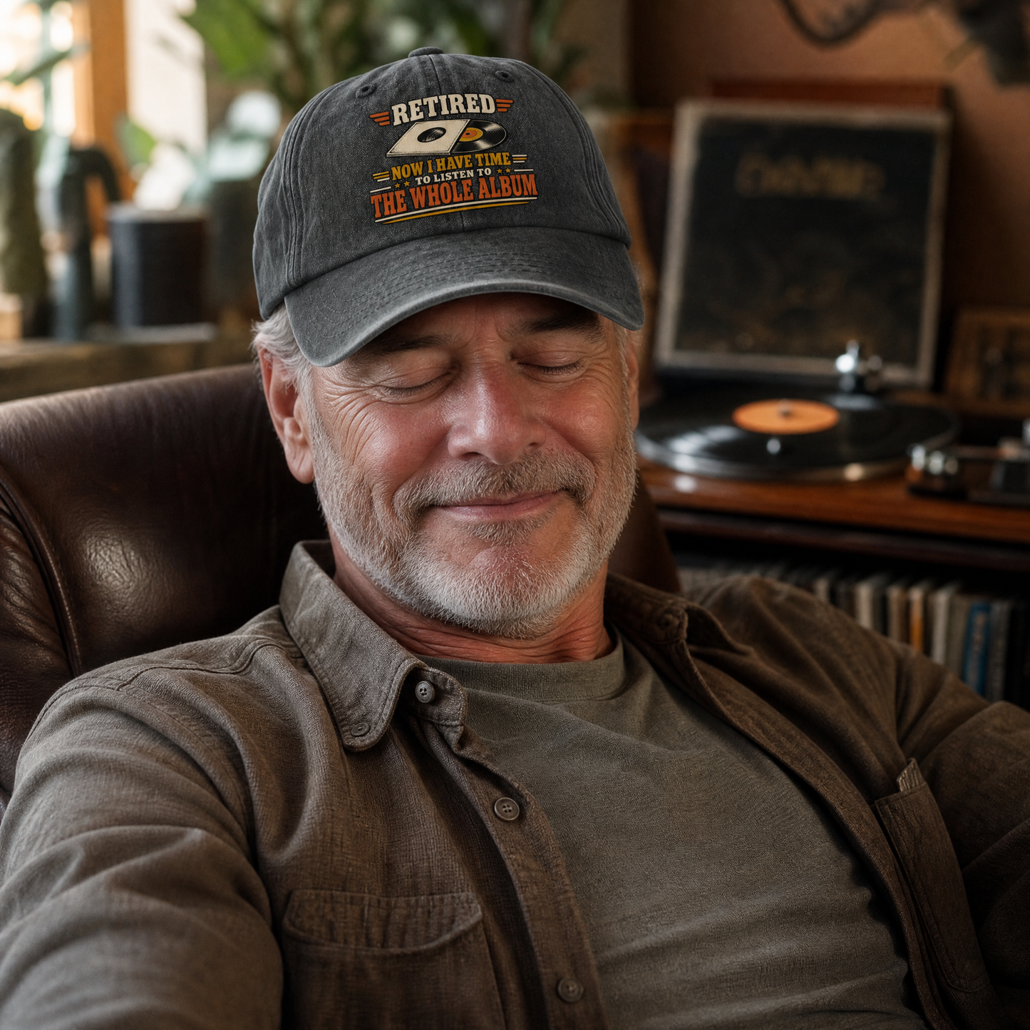 A man wearing a dark gray baseball cap with a graphic that reads 'RETIRED NOW THAT I HAVE THE WHOLE ALBUM' sits in a brown leather chair in a room with a record player in the background.