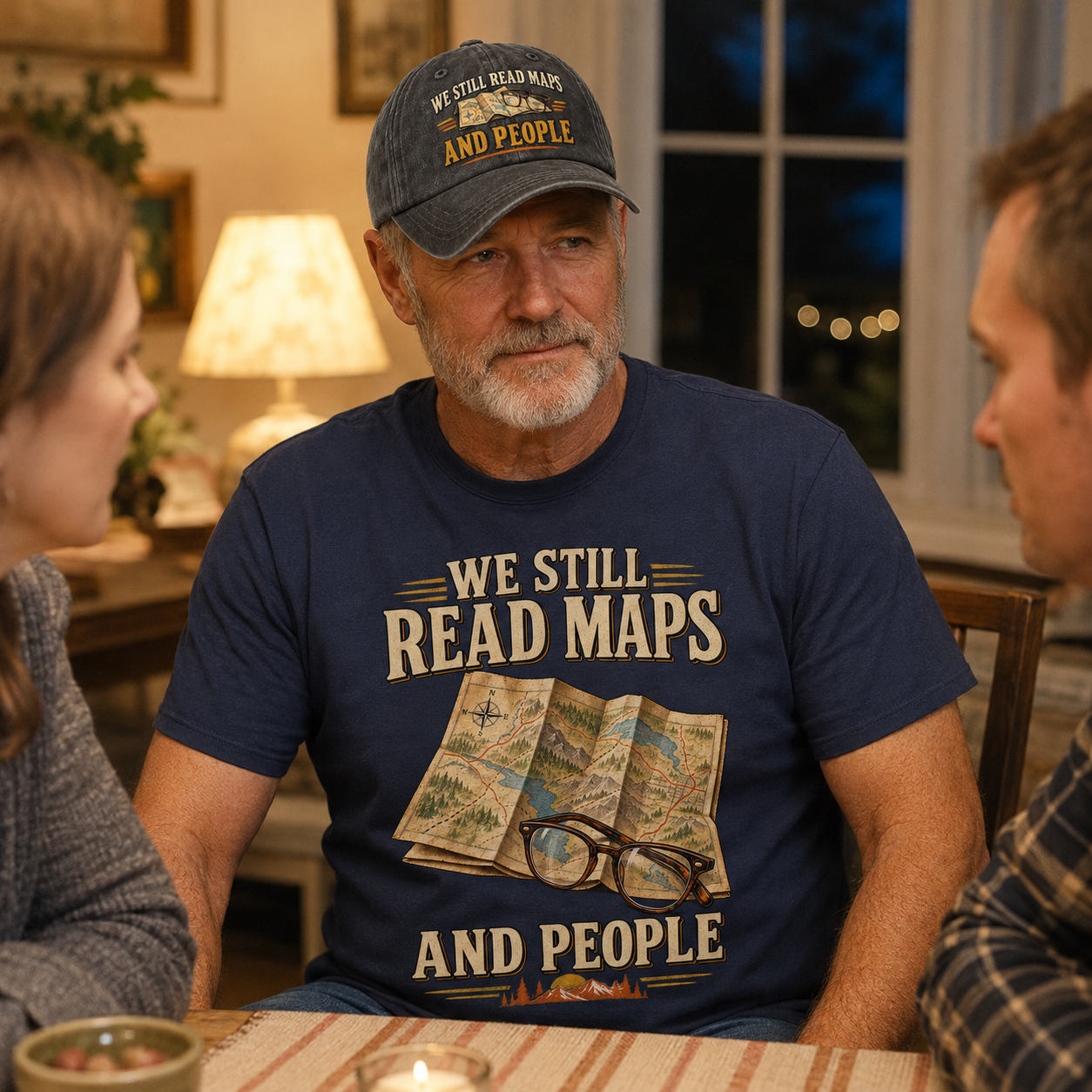 A man wearing a navy blue graphic t-shirt and a matching baseball cap sits at a dinner table with two women, in a warmly lit home setting. The t-shirt features a vintage-style map graphic with the text 'WE STILL READ MAPS AND PEOPLE'.