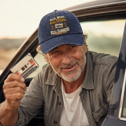 believable older man around 62 with weathered skin, gray stubble, and deep smile lines wearing the "Real Music. Real Friends. Real Summers" vintage washed cap, leaning against the open car door, holding up a cassette, arguing playfully with a friend just off-frame about who picks the song