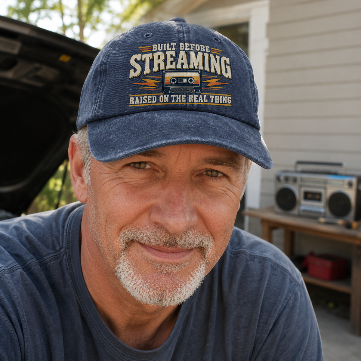 A smiling man wearing a navy blue baseball cap with a retro cassette graphic and the text 'BUILT BEFORE STREAMING RAISED ON THE REAL THING', photographed outdoors in front of a house with a boombox on a table in the background.