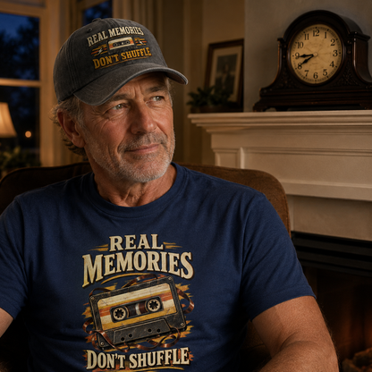 A man wearing a navy blue graphic t-shirt and a dark gray baseball cap, both featuring the text 'REAL MEMORIES DON'T SHUFFLE' and a cassette tape graphic, sitting in a cozy, warmly lit living room.
