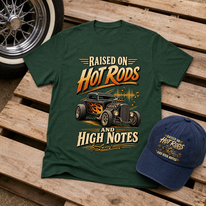 A dark green graphic T-shirt laid flat on a wooden surface, next to a navy blue baseball cap. The shirt features a hot rod car illustration and the text 'RAISED ON Hot RODS AND HIGH NOTES' in orange, yellow, and cream lettering.