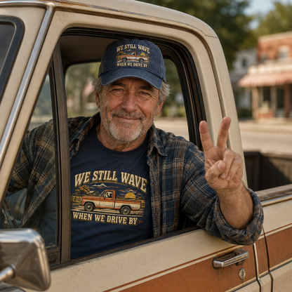 An older man sitting in the driver's seat of a vintage pickup truck, wearing a navy blue graphic t-shirt and matching baseball cap with the text 'WE STILL WAVE WHEN WE DRIVE BY'. He is making a peace sign with his hand, with a blurred outdoor background.