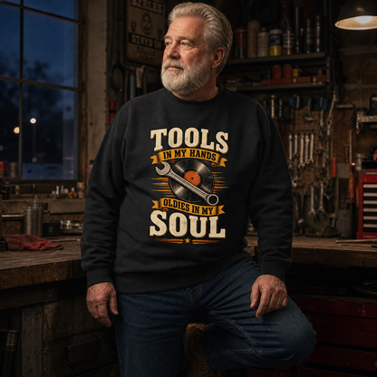 older man in his early 60s with a fuller build and a settled expression wearing the "Tools In My Hands. Oldies In My Soul" vintage sweatshirt as his outer layer, long sleeves clearly visible, on a cool evening, leaning against the workbench with one foot up on a lower shelf