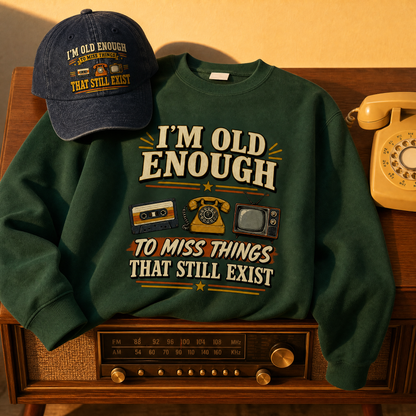 A dark green crewneck sweatshirt with a retro graphic reading 'I'M OLD ENOUGH TO MISS THINGS THAT STILL EXIST' is styled on a wooden surface next to a matching navy baseball cap and a vintage cream rotary phone.