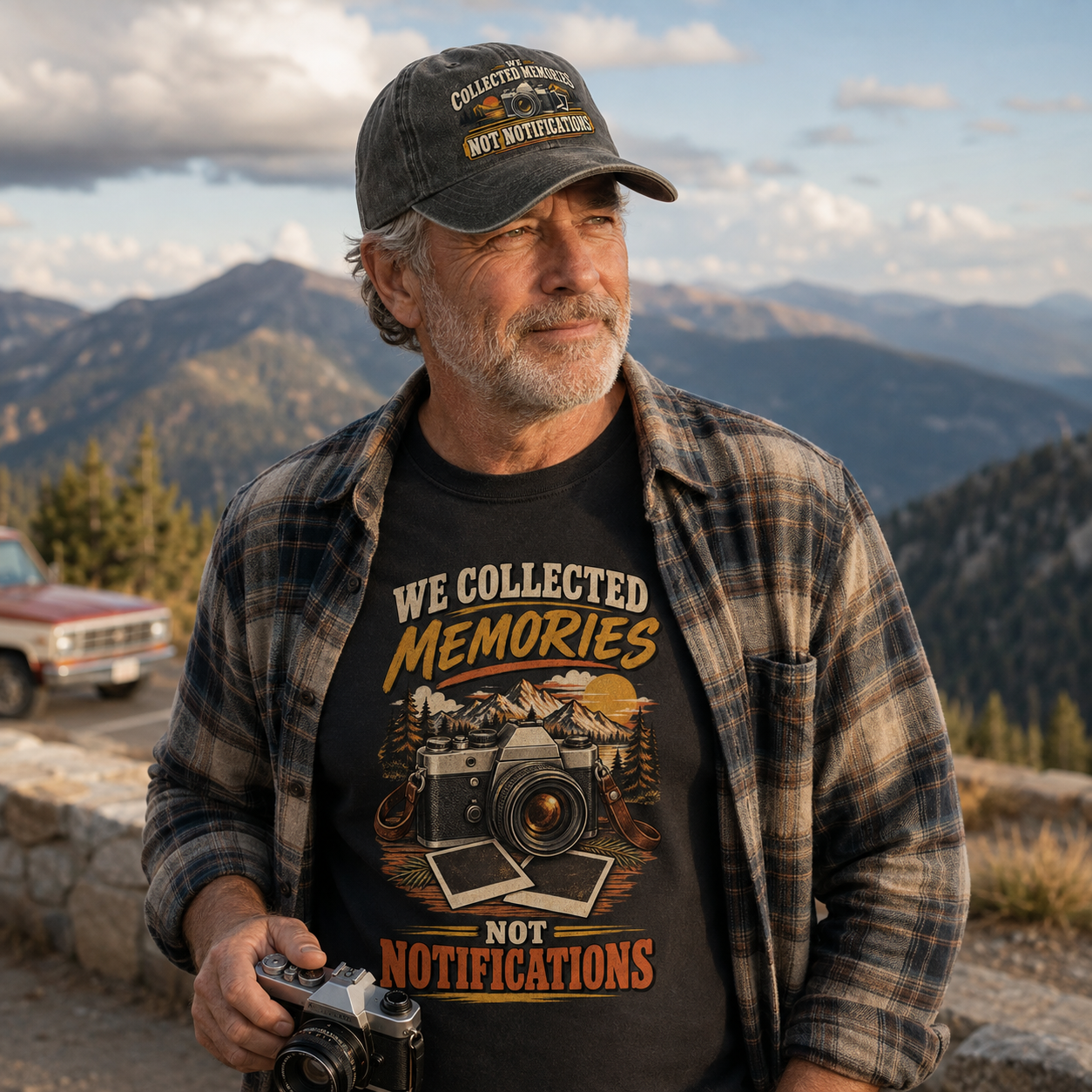 An older man wearing a black graphic t-shirt with a vintage camera and mountain design, a plaid flannel shirt, and a dark baseball cap, holding a camera in front of a mountain landscape.