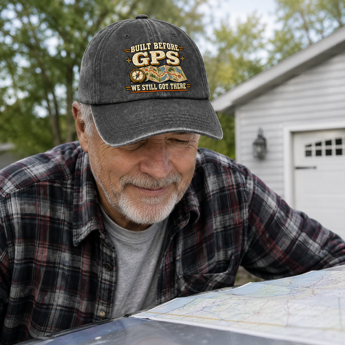 An older man wearing a dark gray baseball cap with a 'Built Before GPS We Still Got There' graphic, leaning over a map in front of a white garage and trees.