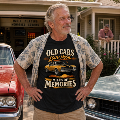 An older man stands outdoors between two classic cars, wearing a black graphic T-shirt with a yellow muscle car print and the text 'OLD CARS LOUD MUSIC MILES OF MEMORIES,' plus a light floral short-sleeve button-up shirt. A house and porch are in the background.