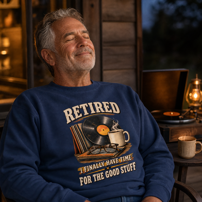 A smiling older man wearing a navy blue crewneck sweatshirt with a graphic that reads 'RETIRED I FINALLY HAVE TIME FOR THE GOOD STUFF' with a vinyl record, glasses, and coffee cup illustration. The scene is set in a cozy, warmly lit rustic cabin interior at night.
