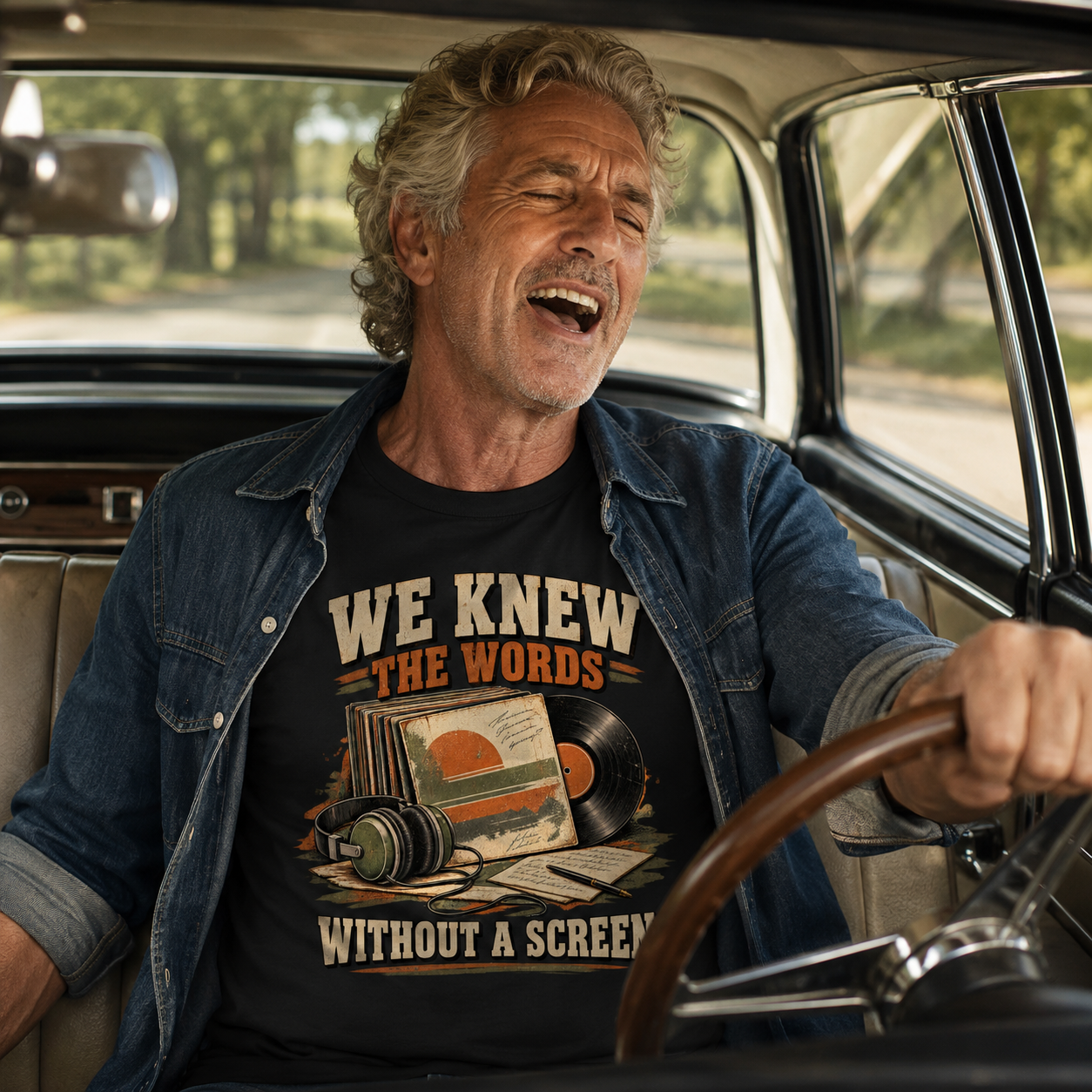 A man laughing while driving a vintage car, wearing a black graphic t-shirt with a retro vinyl record design and the text 'WE KNEW THE WORDS WITHOUT A SCREEN'.