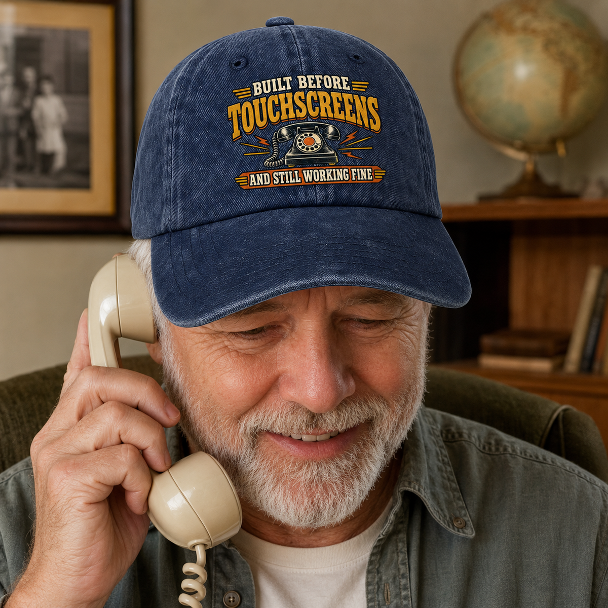 An older man with a gray beard is smiling while talking on a vintage cream-colored telephone. He is wearing a navy blue baseball cap with a distressed yellow and orange graphic that reads 'BUILT BEFORE TOUCHSCREENS AND STILL WORKING FINE'.