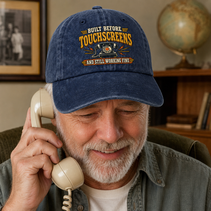 An older man with a gray beard is smiling while talking on a vintage cream-colored telephone. He is wearing a navy blue baseball cap with a distressed yellow and orange graphic that reads 'BUILT BEFORE TOUCHSCREENS AND STILL WORKING FINE'.
