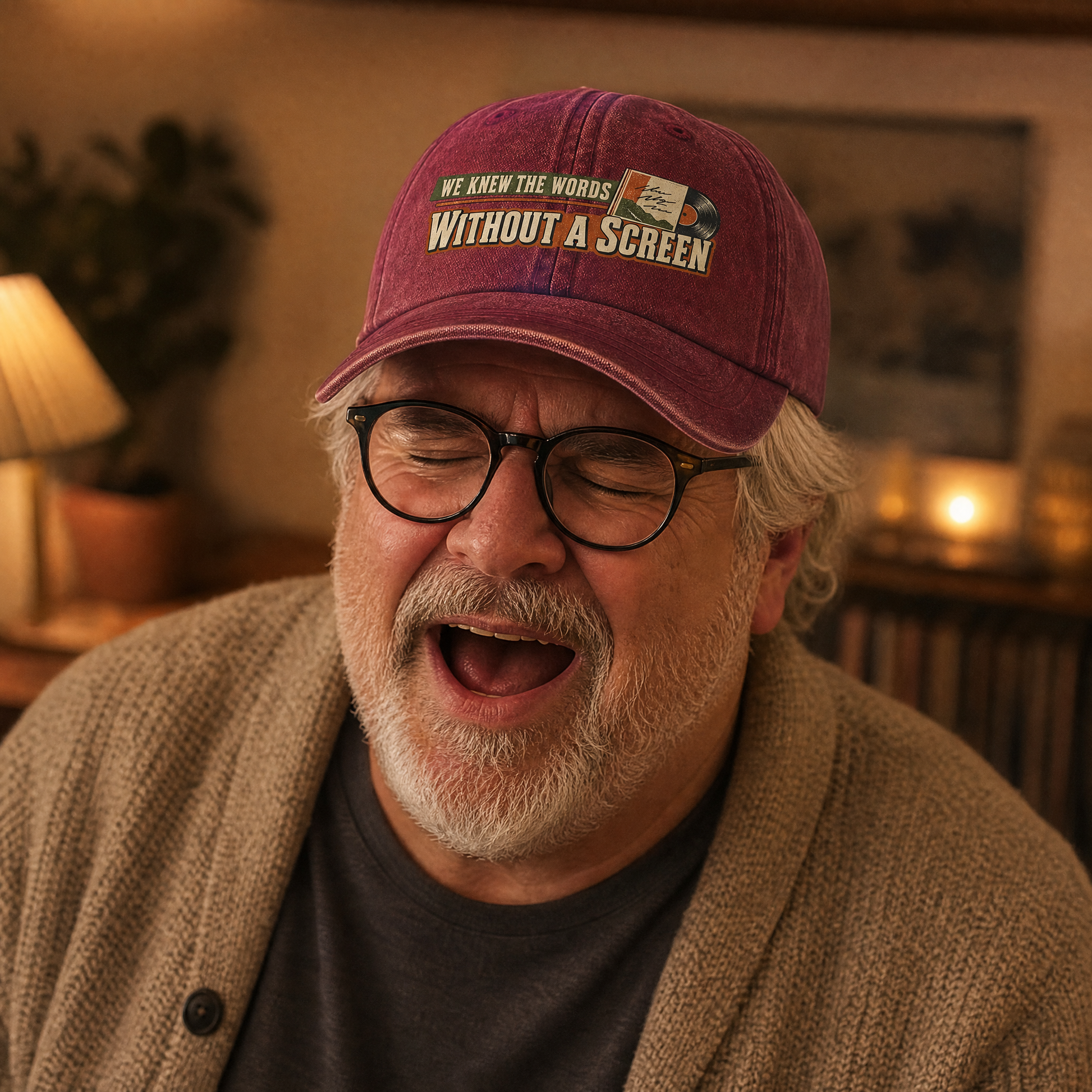 A man wearing a maroon baseball cap with the text 'WE KNEW THE WORDS WITHOUT A SCREEN' and glasses, laughing in a warmly lit indoor setting.