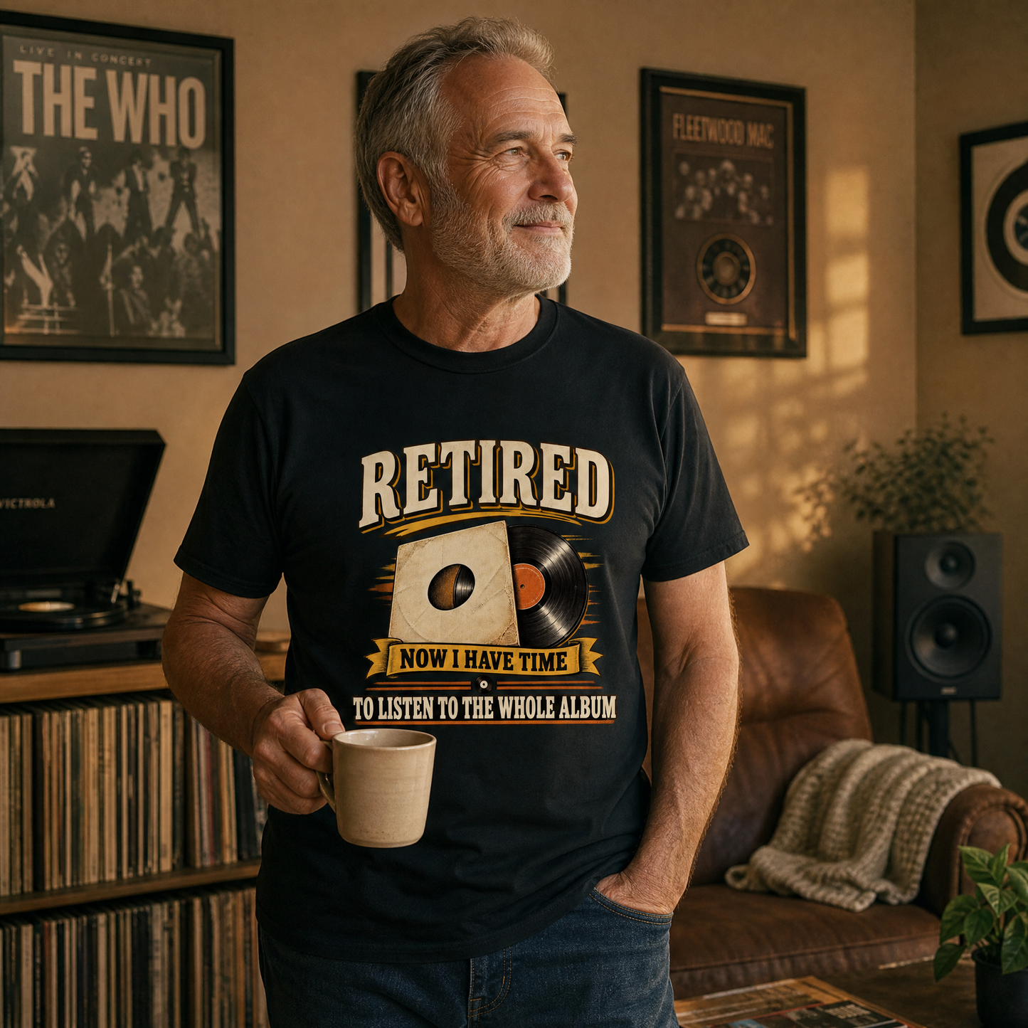 A man in a living room wearing a black graphic t-shirt that reads 'RETIRED NOW I HAVE TIME TO LISTEN TO THE WHOLE ALBUM'. He is holding a mug, with framed Fleetwood Mac posters on the wall behind him.