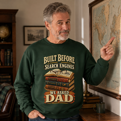 A man wearing a dark green crewneck sweatshirt with a vintage-style graphic that reads 'BUILT BEFORE SEARCH ENGINES WE ASKED DAD' above an illustration of stacked books and glasses. He is standing in a room with bookshelves and a framed world map on the wall.