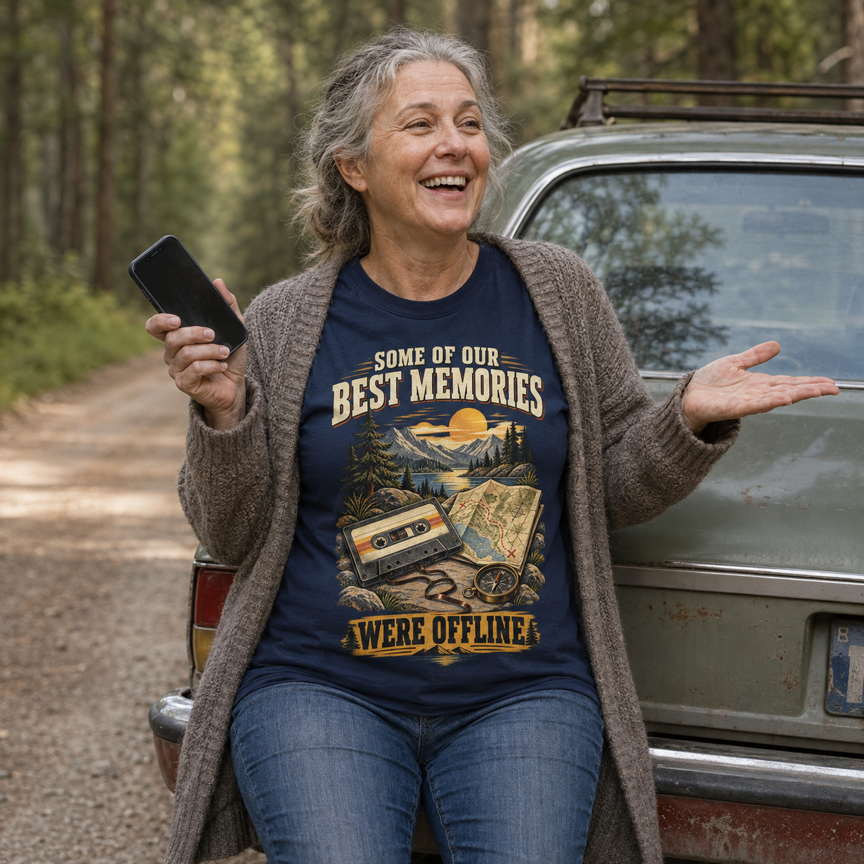 A woman smiling and gesturing while wearing a navy graphic T-shirt with a vintage cassette tape and map design, layered under a brown cardigan, standing beside an old car on a forest road.