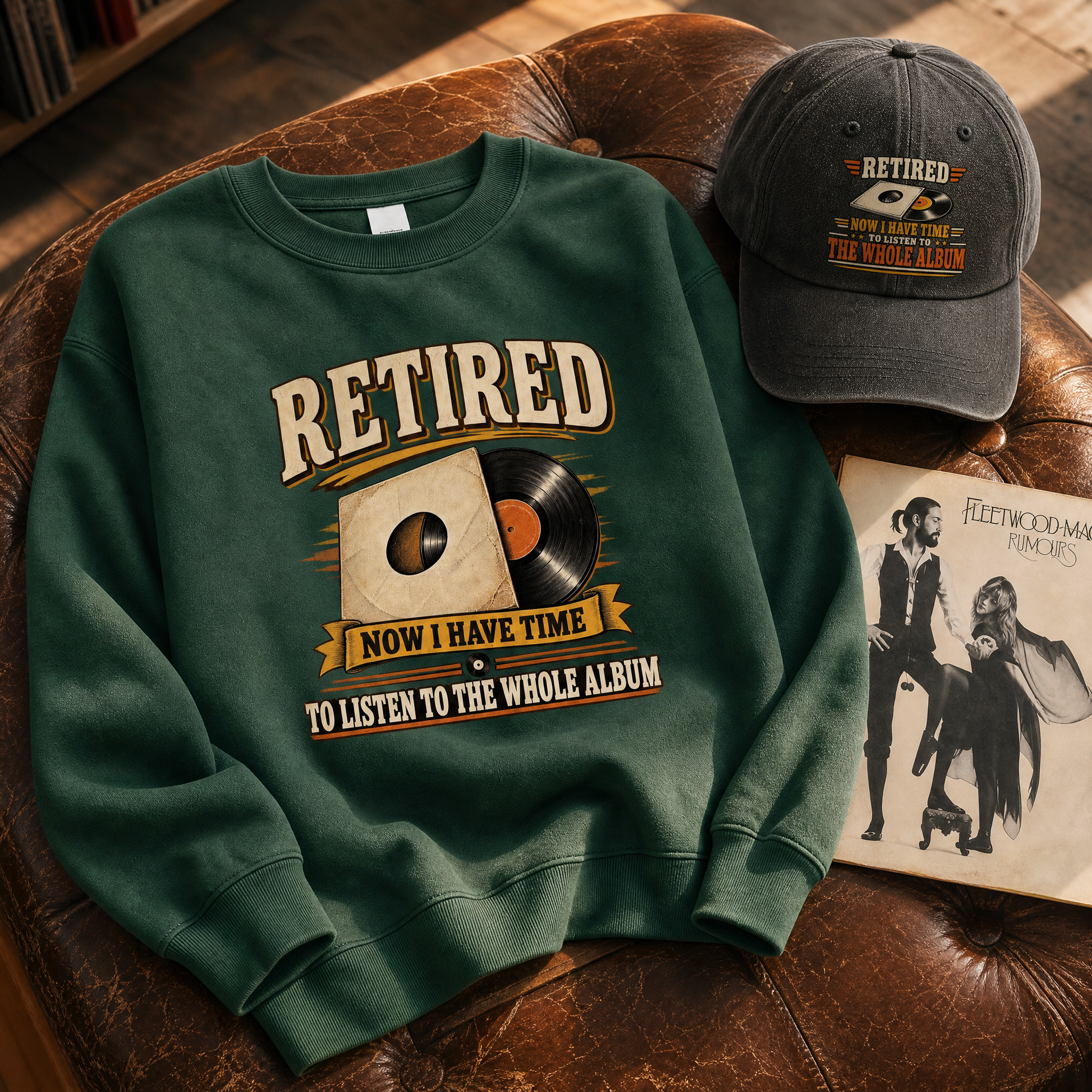 A dark green crewneck sweatshirt with a vintage-style graphic reading 'RETired NOW I HAVE TIME TO LISTEN TO THE WHOLE ALBUM' is styled on a brown leather couch alongside a matching baseball cap and a Fleetwood Mac album cover.