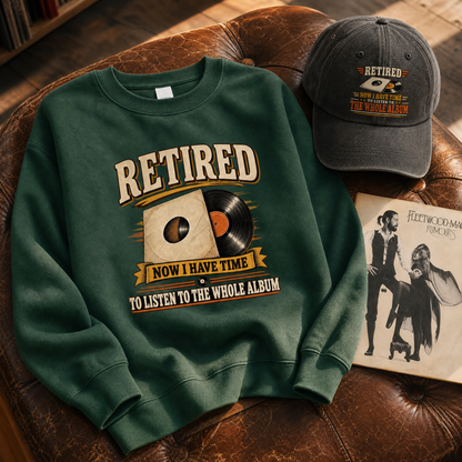 A dark green crewneck sweatshirt with a vintage-style graphic reading 'RETired NOW I HAVE TIME TO LISTEN TO THE WHOLE ALBUM' is styled on a brown leather couch alongside a matching baseball cap and a Fleetwood Mac album cover.