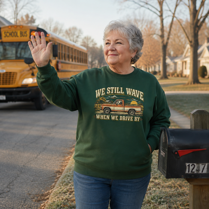 An older woman waving in a residential neighborhood wears a dark green crewneck sweatshirt with a graphic of a pickup truck carrying a Christmas tree and the text 'WE STILL WAVE WHEN WE DRIVE BY'. A yellow school bus is visible in the background on the left.