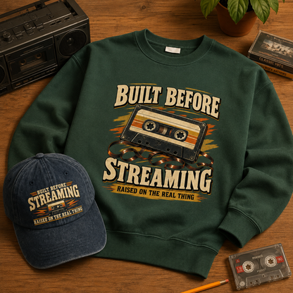 A dark green crewneck sweatshirt with a retro cassette tape graphic and the text 'BUILT BEFORE STREAMING RAISED ON THE REAL THING', shown on a wooden surface alongside a matching navy blue baseball cap, a vintage boombox, a plant, and a cassette tape.