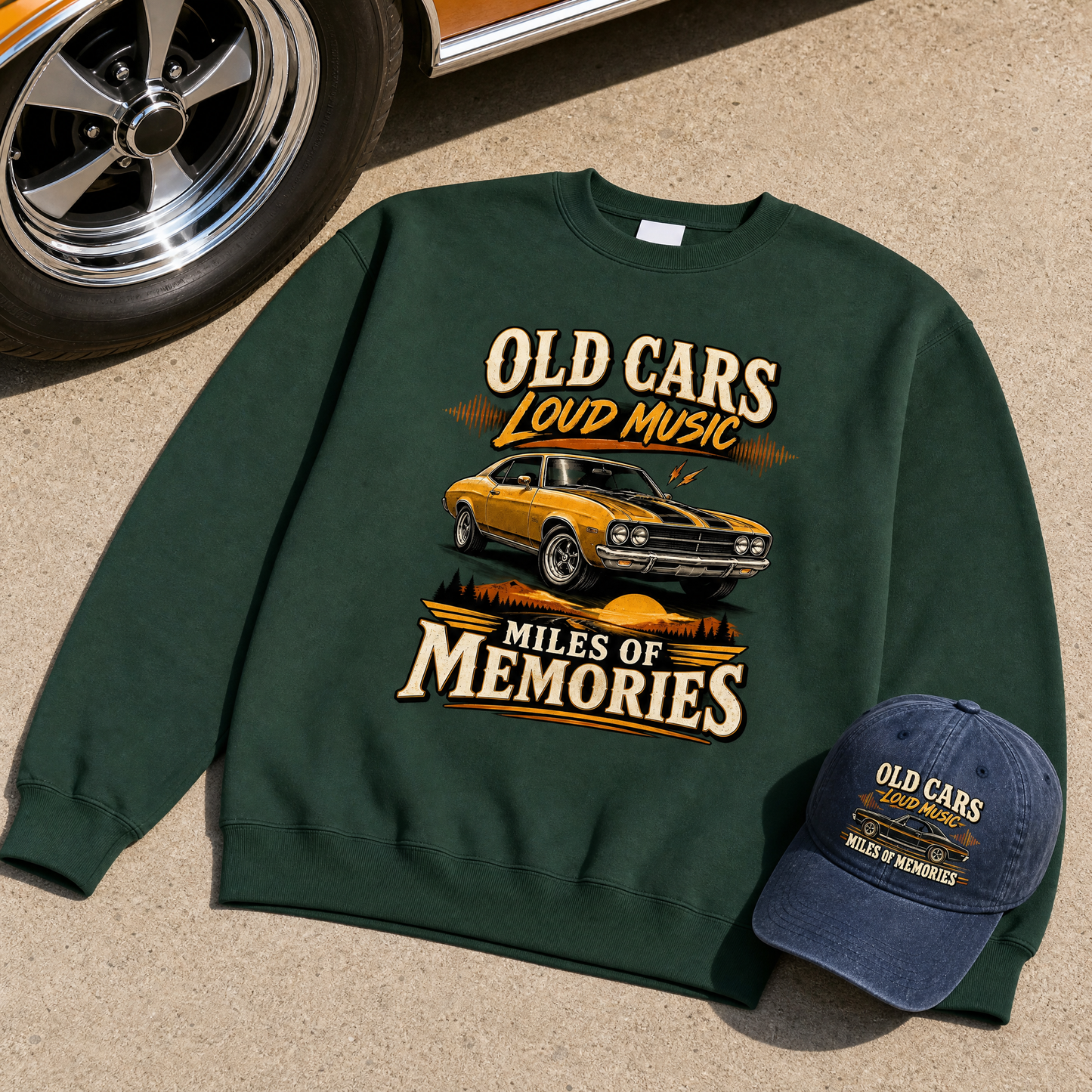 the "Old Cars. Loud Music. Miles Of Memories" vintage sweatshirt and cap as the center of the frame spread flat on the driveway concrete beside a classic car wheel, the fabric lying flat with a soft shadow under it