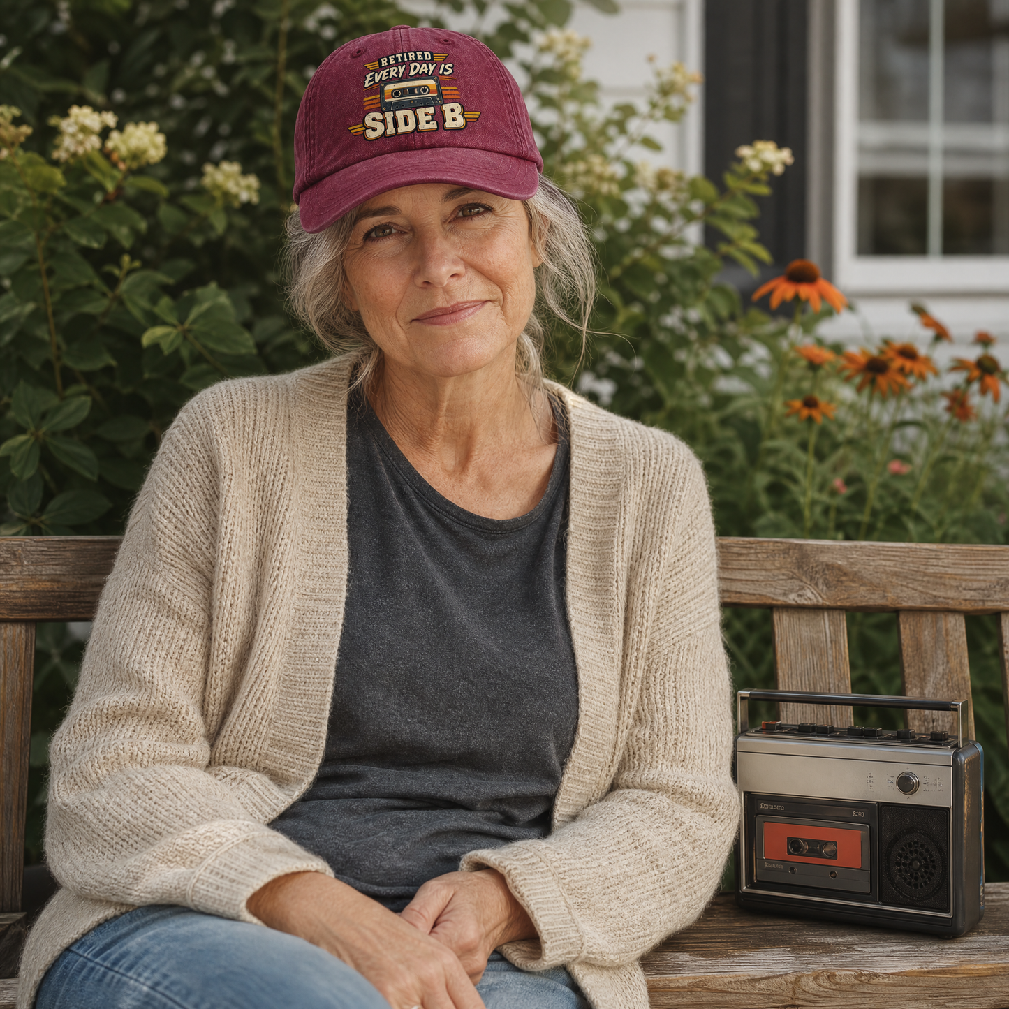 A woman sitting on a wooden bench outdoors, wearing a burgundy baseball cap with 'RIVALRY EVERY DAY IS SIDE B' text, a cream cardigan, and a dark gray t-shirt. A vintage-style boombox sits on the bench next to her.
