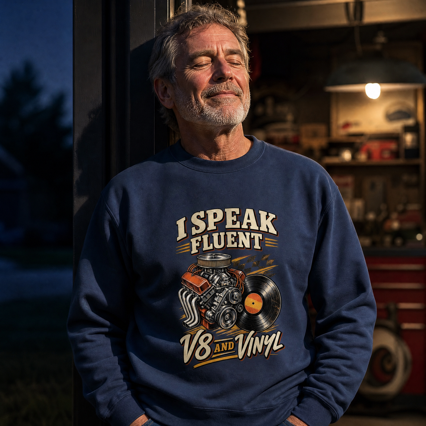 A man wearing a navy blue crewneck sweatshirt with a graphic print that reads 'I SPEAK FLUENT V8 AND VINYL' with an engine and vinyl record illustration, standing in a garage setting.