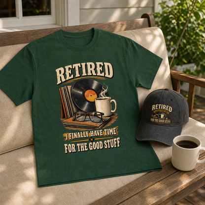A dark green graphic T-shirt laid on a wooden porch chair beside a matching baseball cap, with coffee mugs and a vinyl record illustration on the shirt. The shirt reads “RETIRED I FINALLY HAVE TIME FOR THE GOOD STUFF.